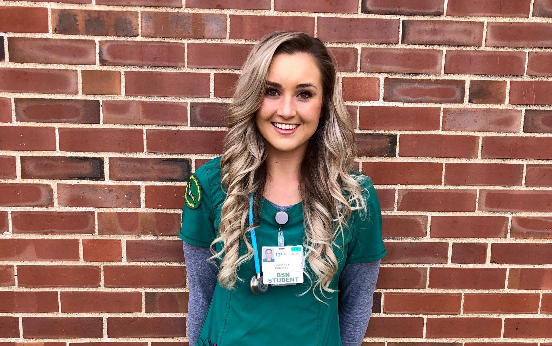 A woman in green scrubs stands in front of a brick wall.